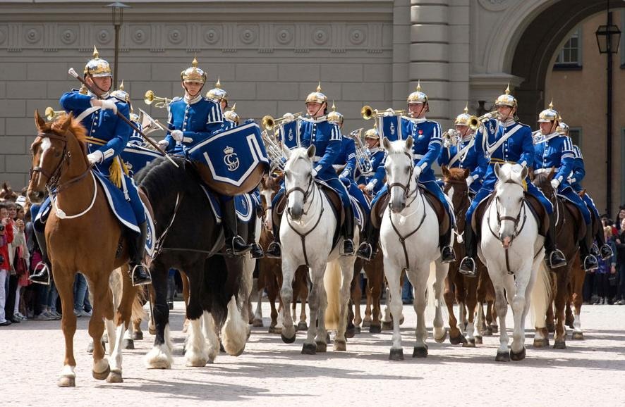 Uniformsklädda ryttare på bruna hästar i rad på slottsgård.