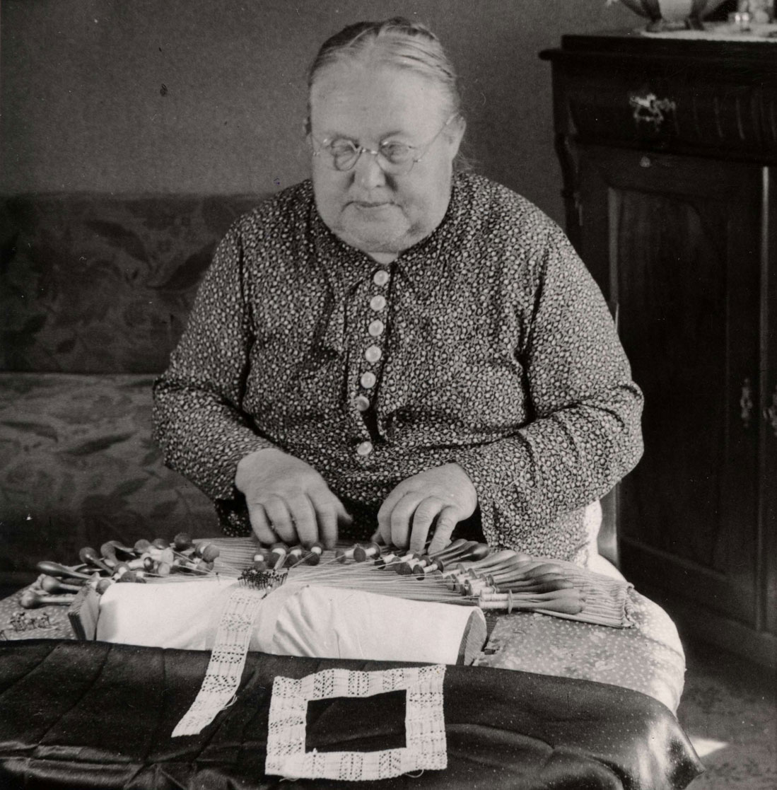 Svartvitt fotografi av kvinna som sitter och arbetar med en spets på en knyppeldyna.