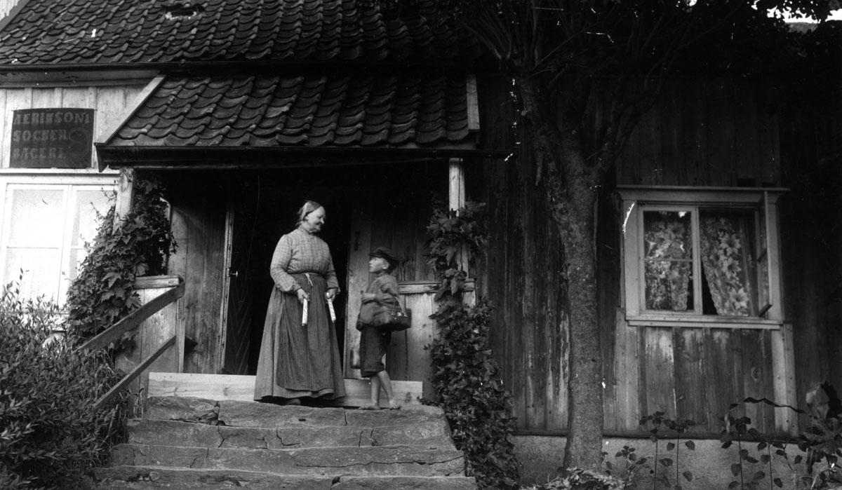 Svartvitt fotografi föreställande kvinna i lång kjol som står på en trapp till ett hus och blickar mot en pojke med korg.