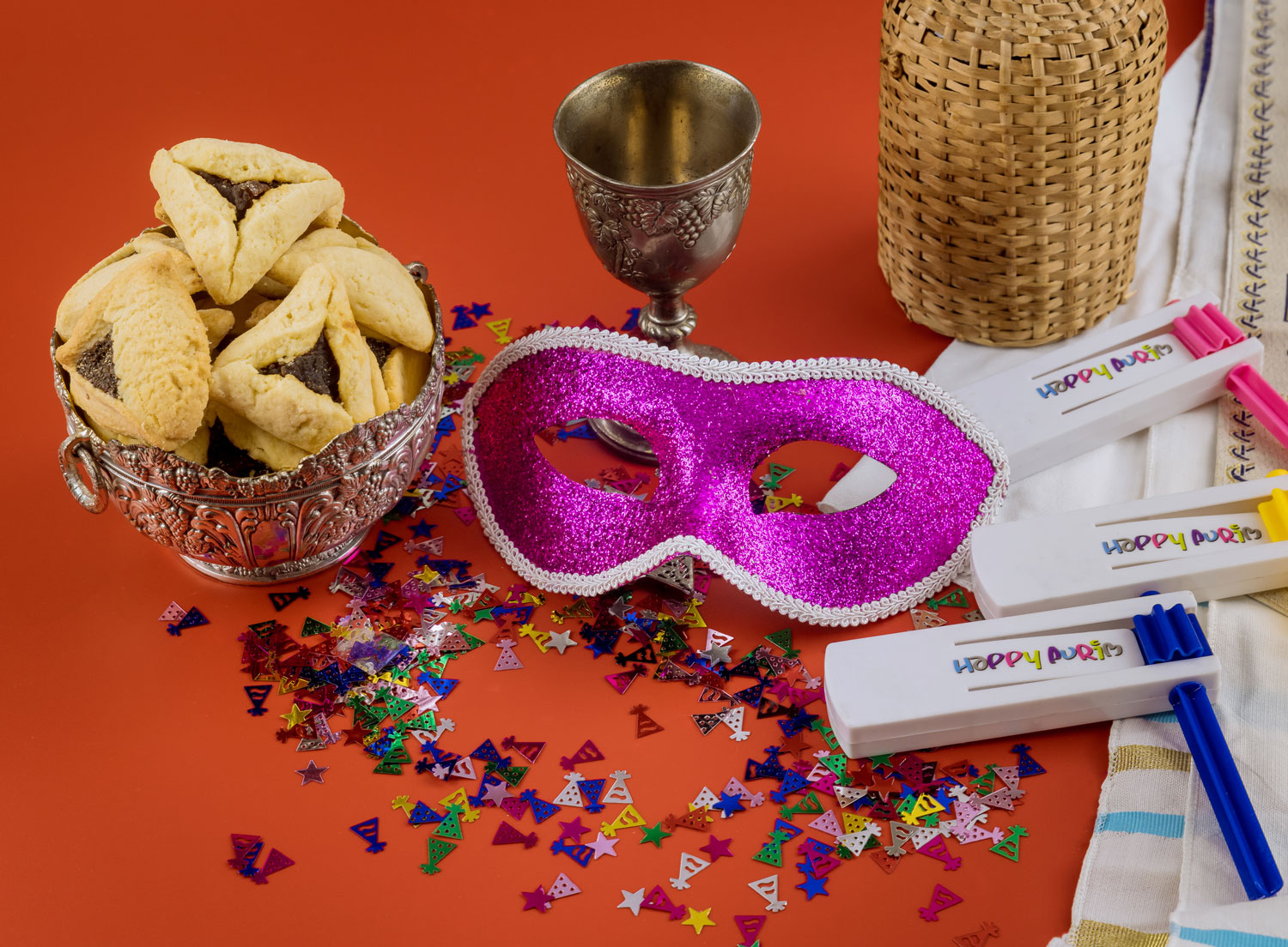 Trekantiga kakor, paljetter, karnevalmasker och skramlor med texten "Happy Purim".