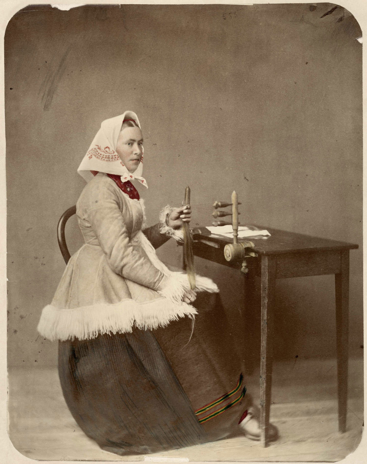 Kvinna i huvudsjal och folkdräkt sitter vid arbetsbord och flätar hår med redskap.