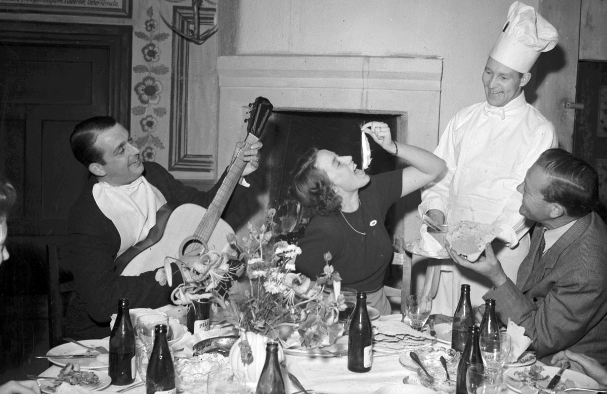 Tre personer runt ett festdukat bord. En kock står vid bordet med fat och en kvinna håller upp en fisk för att stoppa den i munnen.