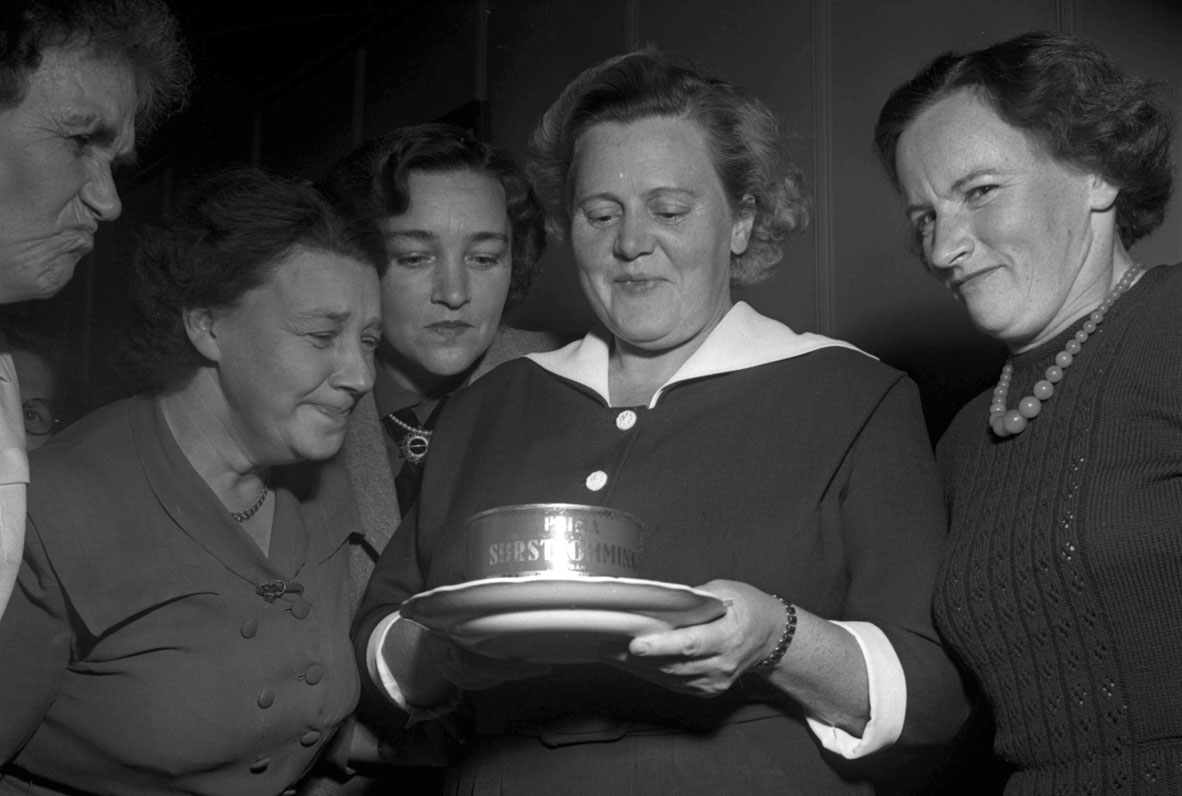Fem kvinnor står runt ett fat med en öppnad surströmmingsburk och ett par av dem grimaserar över lukten.