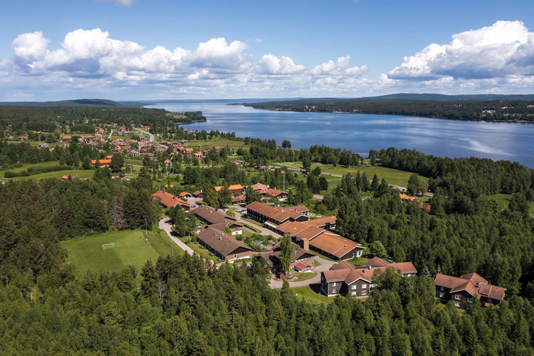 Ett foto taget ovanifrån visar en samling hus som är Västanviks folkhögskola . Skolan ligger omgiven av skog och nära en älv.