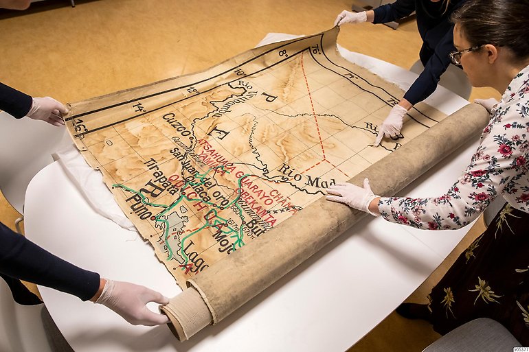 Foto på tre personer som rullar ut en gammal karta på ett bord.